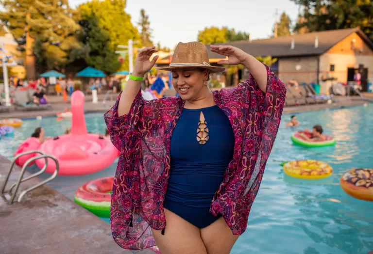 A BIPOC woman in a bathing suit poses in front of a pool filled with floaties.