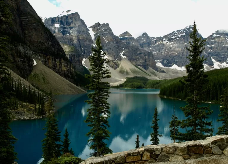 A dark turquoise lake with pine trees in the foreground and mountains in the background.