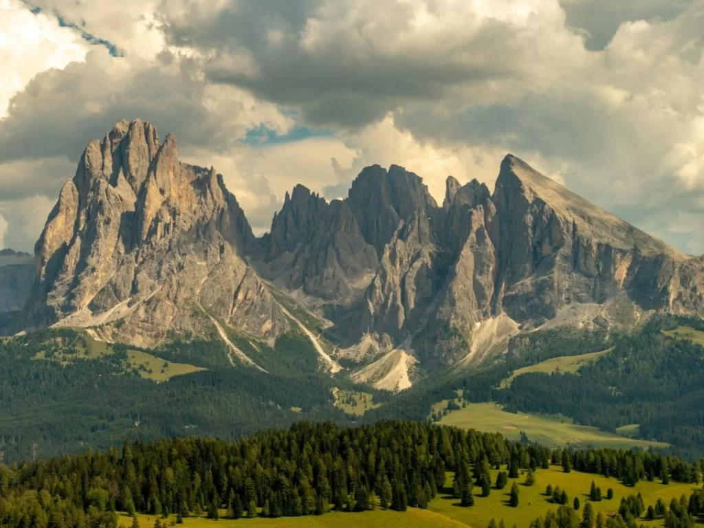 The Dolomites mountains in Italy, with steep rocky mountain peaks, a cloudy sky, and lush green trees and grass at the foot of the mountains. 