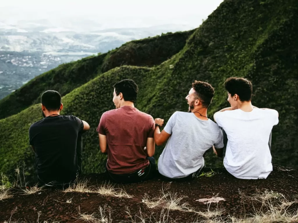 View from behind a group of 4 men laughing and sitting at the top of a grassy hill.