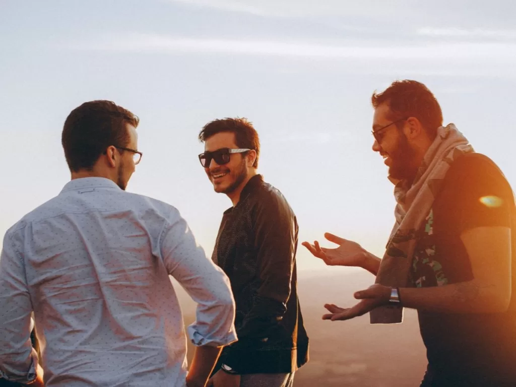 A trio of three men talking and laughing,. Two men are wearing sunglasses and the third is wearing regular eyeglasses. The warm amber lighting in the picture suggests that the sun is setting. 