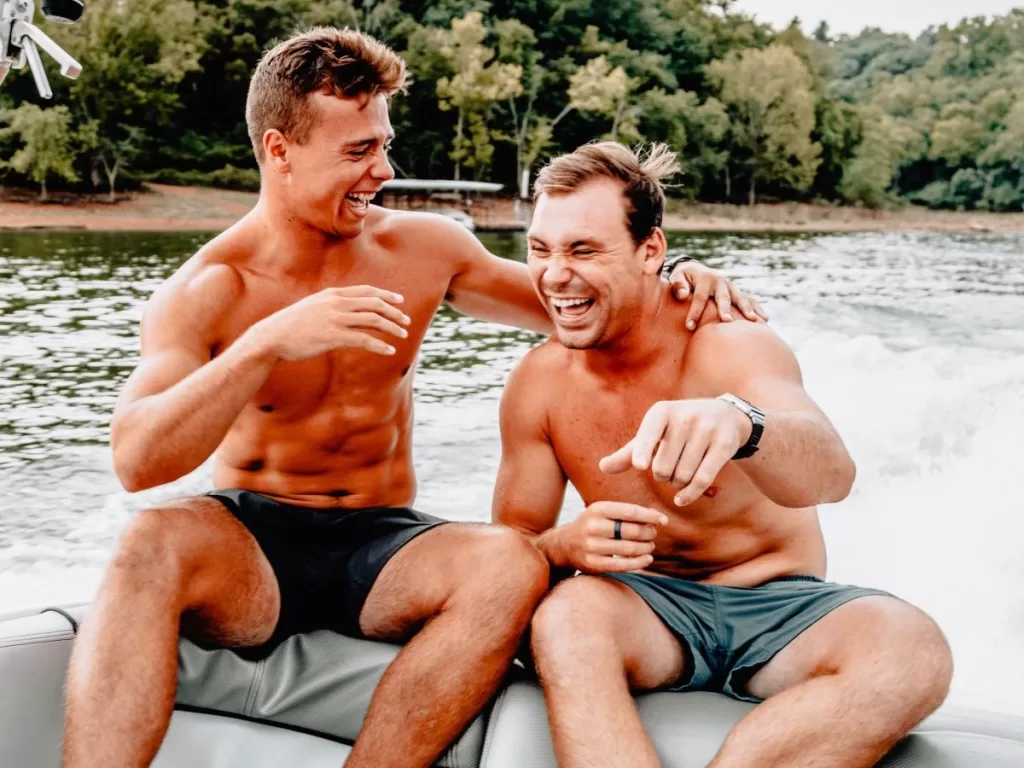Two men laughing and sitting on a boat  with calm water and leafy green trees on the shore behind them.