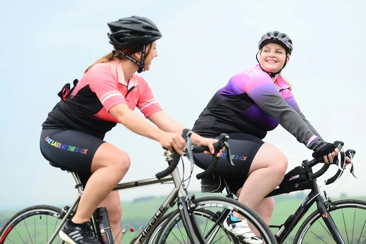 Two bikers riding together wearing pink and purple shirts.