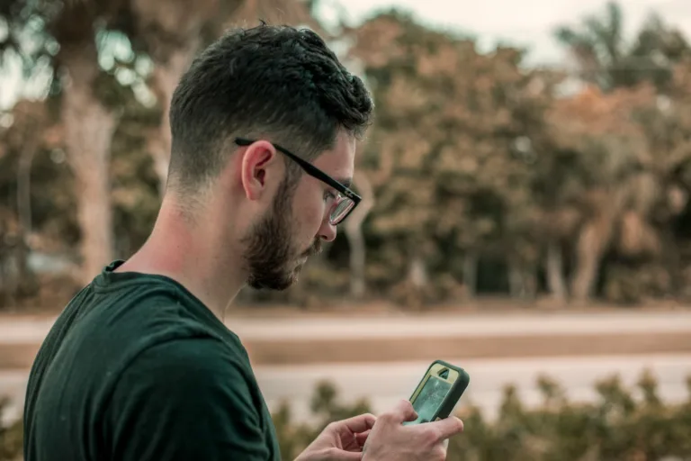 A white man with black glasses, dark hair and a beard looks at a smartphone at the best LGBTQ travel apps.