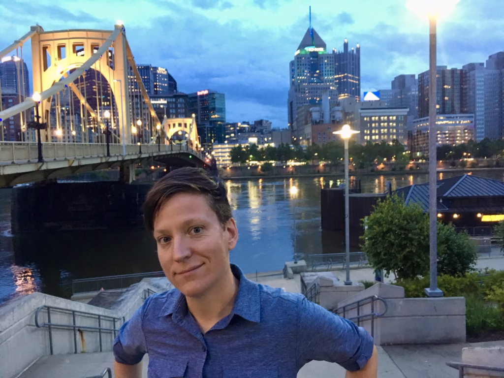A white person in blue shirt standing in front of the river in Pittsburgh at night. 